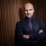 Zdjęcie przedstawia mężczyznę w eleganckim garniturze na drewnianej ścianie. Jego stanowa podstawowa i profesjonalny wygląd może symbolizować sukces i determinację. Fraza „InPost inwestuje” odnosi się do strategicznych działań firmy