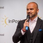 Rafał Brzoska, założyciel firmy InPost podczas konferencji. Ubrany w garnitur, stoi z mikrofonem w ręce i przemawia. Za nim znajduje się baner z logo InPost.
