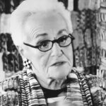 Na zdjęciu znajduje się Rosita Missoni, starsza kobieta o białych włosach. Ubrana we wzorzystą odzież, która pasuje do artystycznego tła. Kobieta nosi okulary o ciemnych oprawkach oraz charakterystyczne kolczyki w szachownicowym wzorze. Wyraz twarzy sugeruje, że opowiada o czymś ważnym. W tle znajdują się kolorowe, abstrakcyjne wzory, które mogą być częścią wystaw, dzieł sztuki lub tekstyliów. Zdjęcie jest wykonane w czerni i bieli, co podkreśla szczegóły i kontrast aplikacji.