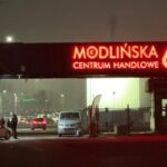 Zdjęcie przedstawia wejście do centrum Modlińska 6D w Warszawie, sfotografowane nocą. Główna brama jest oznaczona znaczenie, podświetlany na czerwono neonowym napisem 'Modlińska 6D Centrum Handlowe'. W tle widać fragmenty parkingu, na które składają się objaśnione samochody, a także kilka osób stojące obok pojazdów, zarysowane na tle ciemnego nieba.