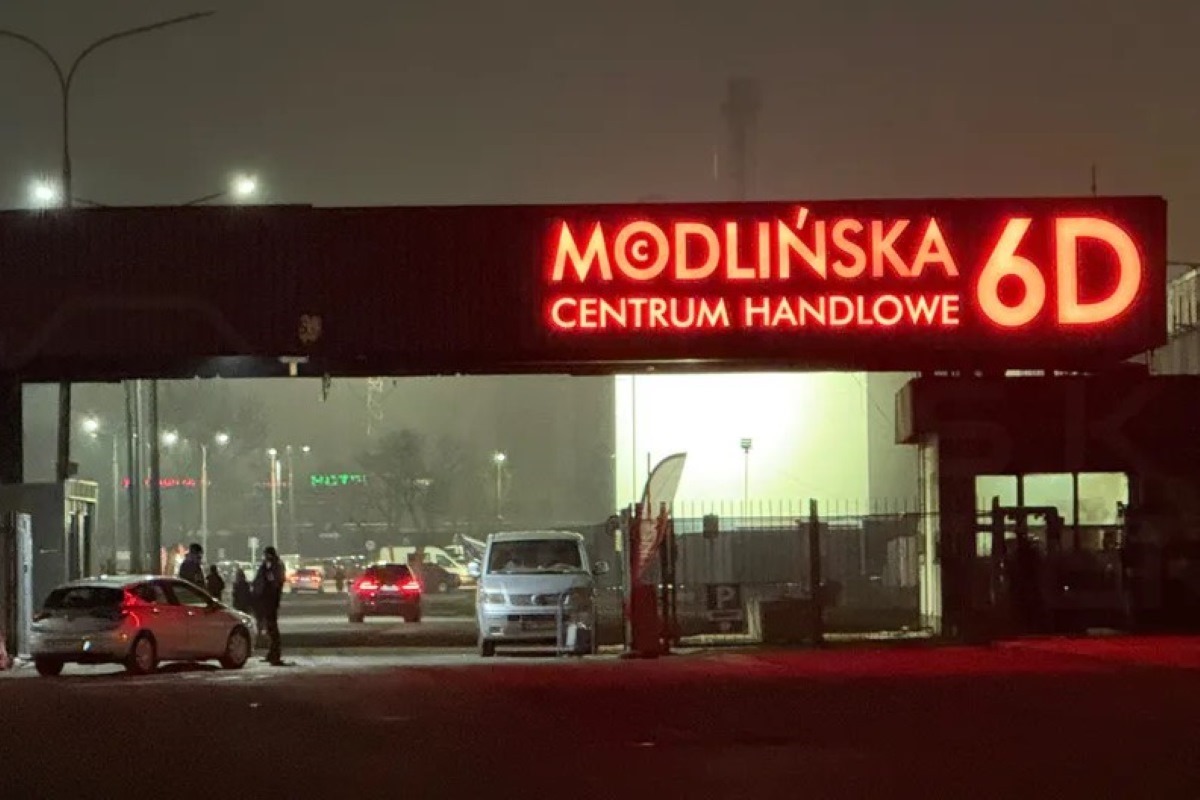 Zdjęcie przedstawia wejście do centrum Modlińska 6D w Warszawie, sfotografowane nocą. Główna brama jest oznaczona znaczenie, podświetlany na czerwono neonowym napisem 'Modlińska 6D Centrum Handlowe'. W tle widać fragmenty parkingu, na które składają się objaśnione samochody, a także kilka osób stojące obok pojazdów, zarysowane na tle ciemnego nieba.