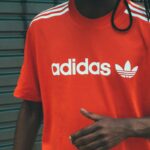 Adidas zapowiada zwolnienia. Na zdjęciu osoba ubrana w czerwony t-shirt marki Adidas z białym logo i napisem 'adidas' na piersi. Koszulka ma charakterystyczne białe paski na ramionach. Tło to metalowa, szara brama z delikatnymi graffiti.