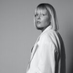 Agnieszka Antosik, CEO The Icon Agency. Nowi klienci dołączają do portfolio agencji. Czarno-białe zdjęcie przedstawiające kobietę z prostymi blond włosami o długości do ramion i grzywce. Ubrana w elegancką marynarkę, patrzy w stronę obiektywu z wyrazem pewności siebie na neutralnym tle.