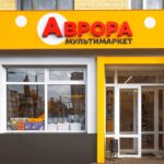Avrora - ukraińska sieć chce zadebiutować w Polsce. Fasada sklepu Avrora w jasnym, żółto-białym kolorze z czerwono-żółtym logo. Na witrynie widoczne są produkty oraz plakaty promocyjne.
