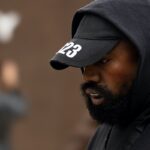 Zdjęcie przedstawia Kanye Westa w czarnej bluzie z kapturem i czapce z napisem '2023', symbolizujące współpracę z marką Adidas, skupione na jego twarzy z poważnym wyrazem, w tle rozmyte postacie.