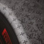Louis Vuitton x Formuła 1. Zbliżenie na oponę z widocznym logo Louis Vuitton wytłoczonym na teksturze powierzchni. Na lewej stronie opony znajduje się czerwony napis 'Pirelli', kontrastujący z ciemnym, chropowatym tłem.