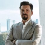 Portret mężczyzny w eleganckim, szarym garniturze, stojącego na tle przeszklonego okna z widokiem na nowoczesną panoramę miasta. Jego profesjonalny wygląd podkreśla zaangażowanie w realizację ambitnych projektów – CCC inwestuje w rozwój i przyszłość swojej sieci logistycznej oraz HalfPrice.