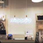 Wnętrze sklepu Moodo – minimalistyczny design z jasnym, neutralnym wystrojem. Na ścianie widnieje podświetlone logo Moodo, a w tle znajdują się różnorodne tekstury dekoracyjne. Po lewej stronie manekiny prezentują ubrania w stylu casualowym, a po prawej akcesoria, takie jak torebki i szale. Centralnie na ladzie kasowej znajdują się urządzenia do obsługi klienta. Całość aranżacji podkreśla nowoczesny i elegancki charakter miejsca.