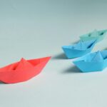 Zdjęcie przedstawia papierowe łódki wykonane techniką origami, ułożone na jasnym tle. Czerwona łódka wyróżnia się na tle niebieskich, symbolizując kreatywność i wyjątkowość. W kontekście "pani od japońskiego" obraz nawiązuje do japońskiej sztuki składania papieru, która jest integralną częścią nauki kultury i języka Japonii.