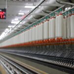 Na zdjęciu widzimy wnętrze nowoczesnej fabryki tekstyliów z długim rzędem maszyn przędzalniczych, które niegdyś produkowały tkaniny dla jednego z najbardziej znanych producentów. Obraz symbolizuje bankructwo popularnego producenta tekstyliów, odzwierciedlając koniec ery przemysłowej tradycji i upadek firmy o wieloletniej historii.