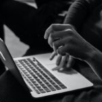Na czarno-białej fotografii widać zbliżenie na osobę korzystającą z laptopa — w centrum kadru znajduje się ręka uniesiona nad klawiaturą, jakby w trakcie wykonywania gestu lub pisania. Laptop jest otwarty, a tło jest nieostre i ciemne, podkreślające sylwetkę użytkownika. Oszustwo na zwrotach pod Zgierzem.