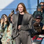 Stella McCartney wykupuje udziały od LVMH i odzyskuje kontrolę nad firmą. Projektantka w eleganckim oversize'owym garniturze na pokazie mody, otoczona publicznością, z której wiele osób robi zdjęcia telefonami.