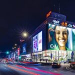 Calzedonia znika z Marszałkowskiej. Zdjęcie przedstawia nocny widok na budynek handlowy Wars Sawa Junior w Warszawie, z dużymi, jasnymi reklamami na fasadzie, w tym C&A, H&M oraz TK Maxx. W tle widoczne są inne budynki miasta, oświetlone światłami miejskimi. Na ulicy przed budynkiem widoczne są światła samochodów w ruchu, tworzące świetliste smugi.