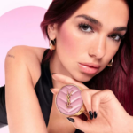 Dua Lipa trzymająca róż do policzków marki Yves Saint Laurent w delikatnym różowym odcieniu, z złotymi akcentami. Ma subtelny makijaż z różem na policzkach i błyszczącymi ustami, ubrana w czarny top i złote kolczyki w kształcie kół, pozująca na tle w pastelowych odcieniach różu i bieli.