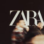Zara na Marszałkowskiej - co zajmie jej miejsce? Logo sklepu Zara na ciemnym tle, w centrum kadru. W tle rozmyci przechodnie, jeden mężczyzna na pierwszym planie patrzy na telefon, tworząc dynamiczną atmosferę miejskiego życia.