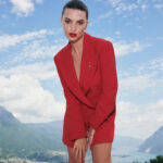 Emily Ratajkowski w kampanii Marella w eleganckiej, czerwonej marynarce stoi pochylona na tle malowniczego krajobrazu gór i błękitnego nieba, z włosami zaczesanymi do tyłu i wyrazistą czerwoną szminką na ustach.