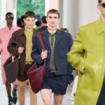 Kering wyniki finansowe Q4 2024. Modele prezentują kolekcję męską podczas pokazu mody Gucci. Ubrani są w eleganckie stylizacje, łączące krótkie spodenki z marynarkami, skórzane płaszcze i modne dodatki, takie jak torby na ramię i chokery. Paleta kolorystyczna obejmuje oliwkową zieleń, granat, róż i brązy.