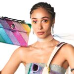 Steve Madden kupuje brytyjską markę Kurt Geiger. Na zdjęciu widać młodą kobietę z warkoczykami i piegami, ubraną w srebrny top bez ramiączek. Trzyma przy twarzy torebkę w intensywnych tęczowych barwach z metalicznym połyskiem Kurt Geiger.
