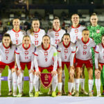 Lancerto ubierze piłkarki na Euro 2025. Reprezentacja Polski w piłce nożnej kobiet pozuje do drużynowego zdjęcia przed meczem. Zawodniczki ubrane są w białe koszulki z czerwonymi akcentami, czerwone spodenki i białe getry. W centrum znajduje się kapitan drużyny trzymająca proporczyk z logo PZPN.