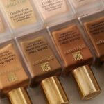 Estee Lauder szykuje masowe zwolnienia. Rząd prostokątnych butelek podkładu Estee Lauder Double Wear w różnych odcieniach (od jasnych po ciemniejsze), z charakterystycznymi złotymi nakrętkami i logo marki na froncie.