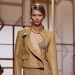 Hermes haute couture. Modelka na wybiegu Hermes prezentująca elegancki, beżowy komplet składający się z asymetrycznej skórzanej kurtki z zamkiem oraz dopasowanego topu i spódnicy.