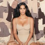 Nike i Skims tworzą nową markę NikeSKIMS. Na zdjęciu widać siedzącą Kim Karsashian o długich, ciemnych włosach, ubraną w dopasowane beżowe body. W tle znajdują się liczne ubrania w odcieniach beżu i brązu.