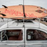Quiksilver, Billabong i Volcom zamykają sklepy. Zdjęcie jasnego, klasycznego samochodu z deską surfingową marki Billabong przymocowaną do bagażnika dachowego. W tle widać zaparkowane pojazdy i osoby na parkingu, a także fragmenty drzew i budynków.