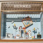 Hermès wyniki finansowe 2024. Witryna sklepu Hermès ozdobiona dużymi kostkami domina, kolorowymi figurkami pionków szachowych, pomarańczowo-niebieskim jedwabnym szalikiem w centralnej części oraz czarną torbą po prawej stronie; aranżacja przypomina motyw gier towarzyskich.