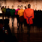 Modowa krzyżówka na Fashion Biznes. Na zdjęciu modelki prezentujące kolekcję na wybiegu Saint Laurent, ubrane w intensywnie kolorowe stylizacje, tworzące modową krzyżówkę barw i fasonów. Na tle eleganckiego, marmurowego tła z logo YSL, każda z sylwetek ukazuje wyrazisty, geometryczny krój i głębokie, nasycone kolory.