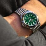 Gallet kupione przez Breitling. Zdjęcie przedstawia zbliżenie na zegarek na męskiej ręce. Zegarek ma srebrną bransoletkę i zieloną tarczę, godzina wskazuje 10:11.