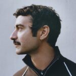 Kim jest Dario Vitale? Zdjęcie przedstawia nowego dyrektora kreatywnego Versace, Dario Vitale. Projektant pozuje bokiem, ma ciemne włosy, kolczyk w uchu i brązową bluzę ze stójką, rozpiętą tuż przy szyi.