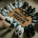 Na zdjęciu widoczne są nogi mężczyzny siedzącego na dywanie. Wokół niego, w kształcie okręgu, ułożone są buty sportowe marki Adidas Yeezy w różnych kolorach i modelach, w tym modele Yeezy Boost 350 i Yeezy 700. W centrum okręgu znajduje się okrągły dywanik z napisem "PLEASE REMOVE YOUR YEEZYS". Mężczyzna ma na sobie czarne szorty i szare skarpety. Podłoga jest jasna, a dywanik beżowy z czarnym napisem.