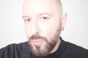 Demna odchodzi z Balenciagi, by zostać nowym dyrektorem kreatywnym Gucci. Na zdjęciu zbliżenie na twarz projektanta z brodą i kolczykiem w uchu, ubranym w czarną koszulkę, na jasnym tle.
