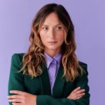 Ekspercka wiedza od specjalistów branży mody, PR, marketingu. Kogo obserwować? Na zdjęciu jedna z ekspertek, Martyna Zastawna. Kobieta z długimi, falującymi włosami w odcieniach brązu z jasnymi końcówkami, ubrana w zieloną marynarkę i fioletową koszulę. Stoi na tle jasnofioletowej ściany z rękami skrzyżowanymi na piersi, patrząc lekko w bok.