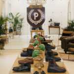 Wnętrze eleganckiego showroomu Pollini z klasycznym wystrojem – skórzane fotele, zielone lampki bankierskie i ekspozycja męskiego obuwia na drewnianym stole.