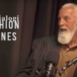 Robert Kupisz ubrany w brązową koszulkę polo i ciemną marynarkę w prążki, siedzi przed mikrofonem w studio nagraniowym. Po lewej stronie widoczny jest napis „dialogi FASHION BIZNES” na tle ciemnych zasłon.