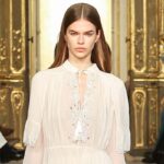 Modelka w białej, romantycznej sukni z delikatnymi haftami, idąca po złotym wybiegu podczas pokazu Twinset na Milan Fashion Week.