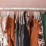 Gerry Weber upadłość kolejnego oddziału. Na zdjęciu wieszak z kolorowymi ubraniami w sklepie odzieżowym Gerry Weber. Ubrania w odcieniach czerni, bieli, pomarańczu i zieleni, z różnorodnymi wzorami, w tym kwiatowymi i abstrakcyjnymi. Wieszak na tle jasnoróżowej ściany.