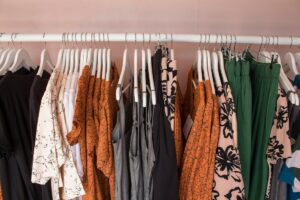 Gerry Weber upadłość kolejnego oddziału. Na zdjęciu wieszak z kolorowymi ubraniami w sklepie odzieżowym Gerry Weber. Ubrania w odcieniach czerni, bieli, pomarańczu i zieleni, z różnorodnymi wzorami, w tym kwiatowymi i abstrakcyjnymi. Wieszak na tle jasnoróżowej ściany.