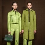 Zdjęcie z backstage'u pokazu Hermès. Dwie modelki pozują na brązowym tle w zielonych total lookach – jedna w jaskrawym odcieniu i torebką w ręce, a druga w długim płaszczu.