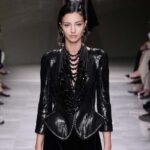 Lucy Markovic. Modelka idzie na wybiegu w trakcie pokazu mody. Ma ciemne włosy i czarny total look. W tle widać widownię.