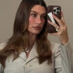 Hailey Bieber robi zdjęcie w lustrze, pozując w beżowej marynarce i rozpuszczonych włosach. Na zdjęciu widać telefon z casem na balsam marki Rhode, która może zostać sprzedana.