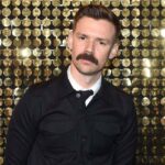 Adam Selman, nowy dyrektor kreatywny Victoria's Secret. Mężczyzna w ciemnych włosach i z wąsem pozuje na złotym tle. Ma na sobie koszulę, krawat i czarną zapiętą kurtkę.