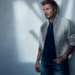 David Beckham w jasnoszarej kurtce bomberce i granatowym T-shircie, oparty o ścianę, prezentuje nowoczesny, miejski look BOSS.