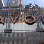 Avon – zwolnienia grupowe. Zdjęcie przedstawia szyld marki Avon na szklanym budynku, w którym odbijają się obiekty po drugiej stronie ulicy.