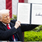 Zdjęcie Donalda Trumpa nakładającego tzw. cła Trumpa na pozostałe kraje na świecie. Prezydent trzyma podpisany dekret, stoi na tle flagi amerykańskiej. Ma na sobie garnitur, czerwony krawat i białą koszulę.