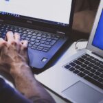 Osoba pracująca na dwóch laptopach – Dell i MacBook – w ciemnym pomieszczeniu, symbolizująca działania związane z cyberbezpieczeństwem i ochroną danych.
