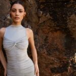 Modelka Chaima Ameziane w jasnej, dopasowanej sukni LA MANIA na tle skalistego wybrzeża, ukazująca harmonię ciała i tkaniny w naturalnym, letnim pejzażu.