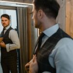 Kluczowe elementy męskiej garderoby - na zdjęciu mężczyzna przeglądający w lustrze, ubrany w elegancką koszulę, kamizelkę i spodnie garniturowe oraz krawat.