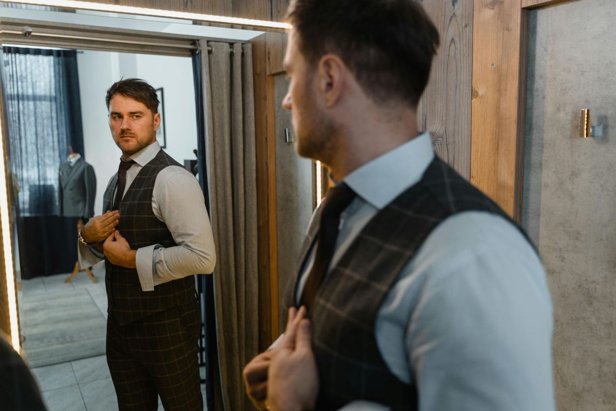 Kluczowe elementy męskiej garderoby - na zdjęciu mężczyzna przeglądający w lustrze, ubrany w elegancką koszulę, kamizelkę i spodnie garniturowe oraz krawat.